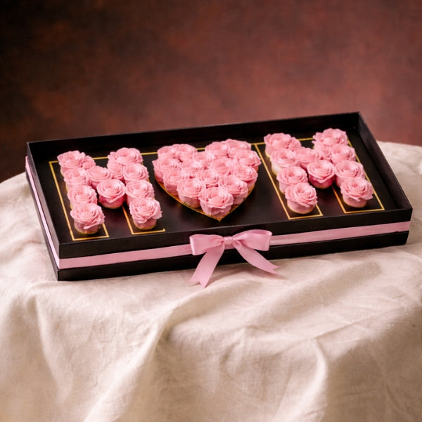 Decorative arrangement of pink roses shaped like the word 'MOM' on a black tray with a pink bow.